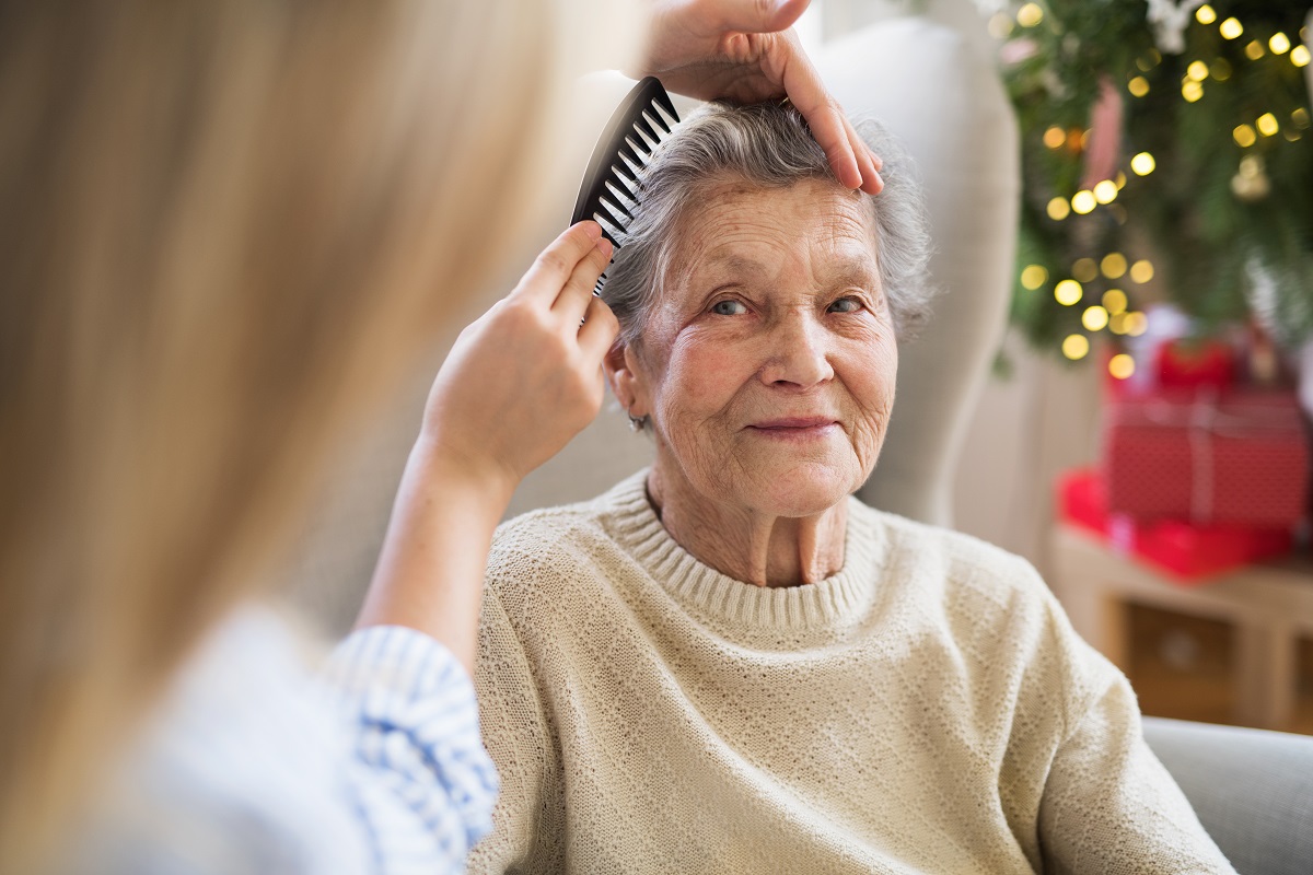 An older woman has her hair combed by a younger woman in front of a decorated Christmas tree.