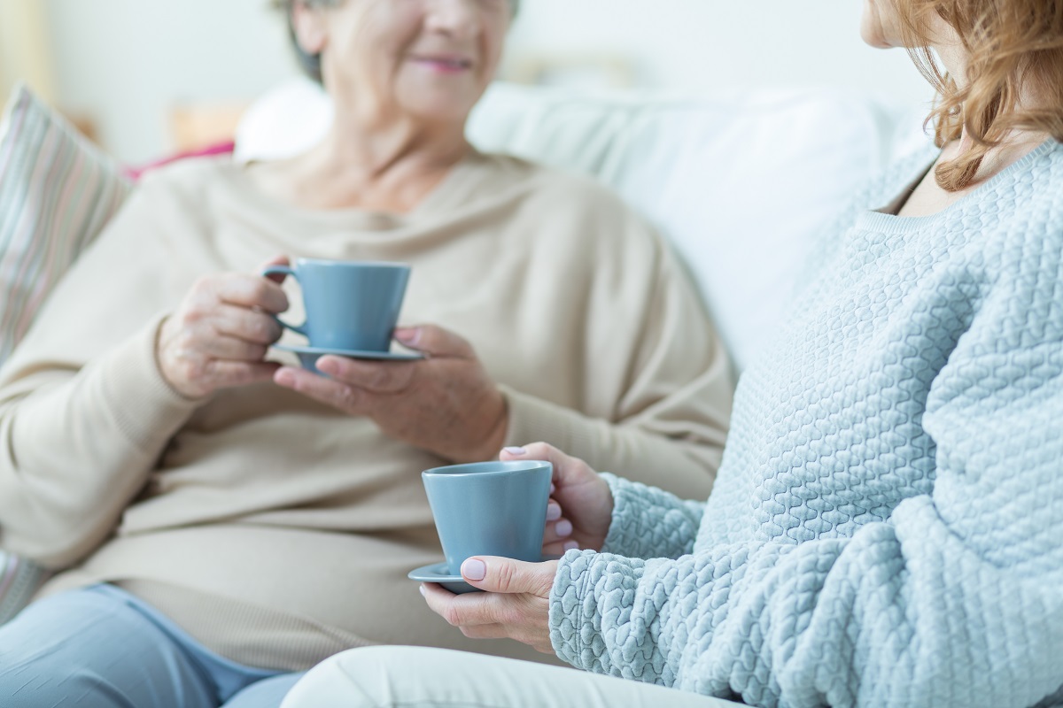 Two women sit together on a sofa holding cups of tea, one comforting the other during recovery.