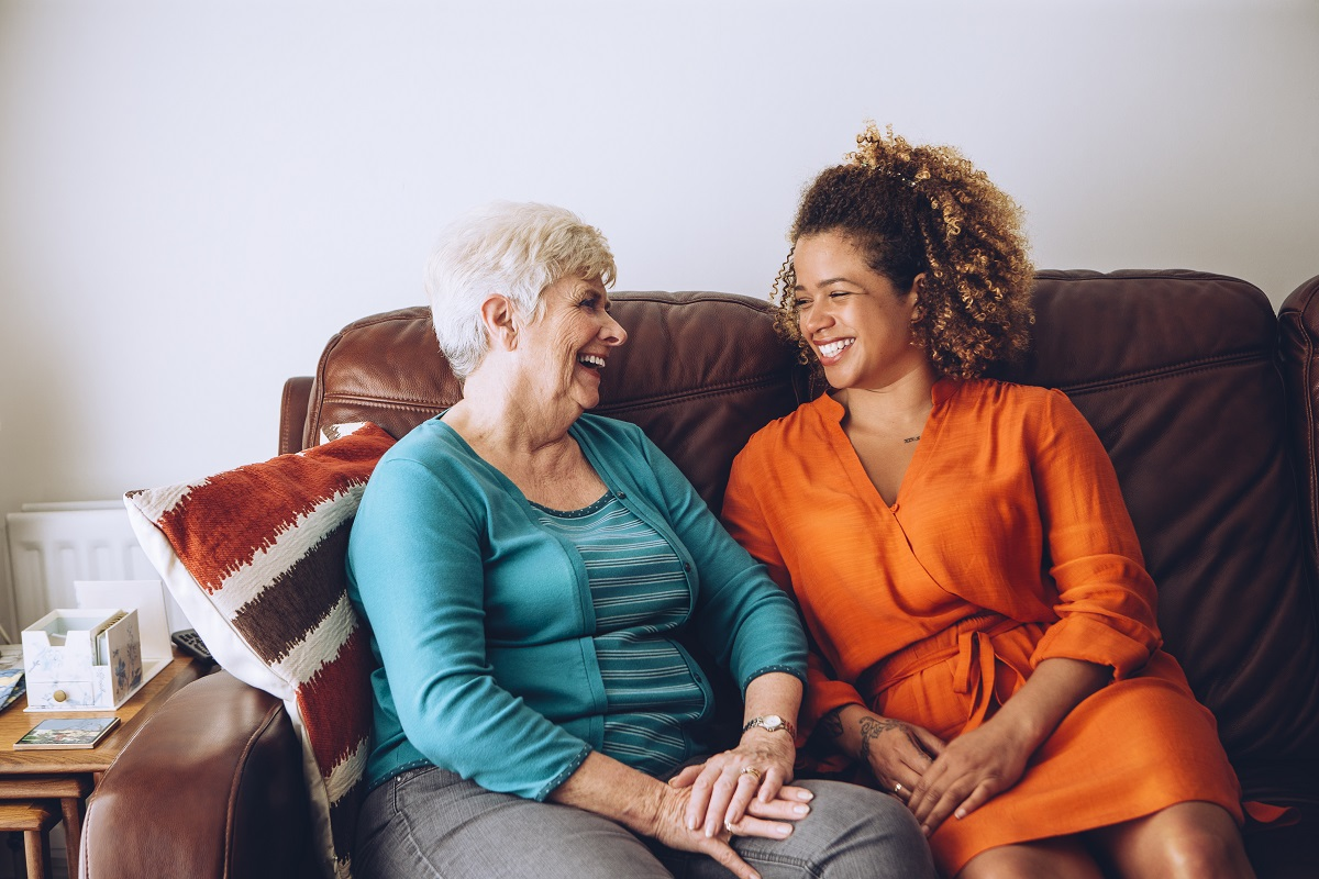 An older woman with white hair and a younger woman with curly hair sit together on a brown leather sofa, smiling and holding hands.